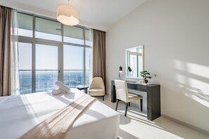 A modern hotel room with a large bed, a desk, a chair, and a view of the city through large windows. - Elegant 2BR in Tower 108 Jumeirah Village Circle by Deluxe Holiday Homes (Dubai)