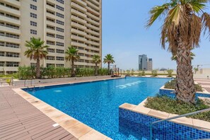A swimming pool with a tiled edge, surrounded by palm trees and a wooden deck. - Elegant 2BR in Tower 108 Jumeirah Village Circle by Deluxe Holiday Homes (Dubai)