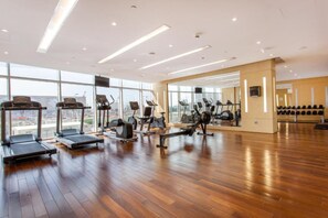 Un gimnasio amplio con diversas máquinas de cardio, grandes ventanales que ofrecen vistas a la ciudad y suelo de madera.