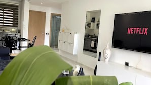 Un salon moderne avec un téléviseur à écran plat fixé au mur, un coin repas avec une table et des chaises, et une grande plante au premier plan.