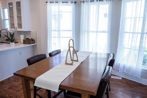 Un comedor con mesa de madera, mantel blanco y sillas. Hay ventanas con cortinas blancas, una zona de cocina con armarios y una planta en maceta sobre la encimera.