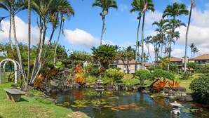 A tropical garden with a pond, palm trees, and a stone bridge. - Pono Kai F207 Oceanview AC, POOL, JACUZZI, BBQ, TENNIS (Kapaa)