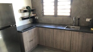 Una cocina con gabinetes de madera, un refrigerador de acero inoxidable, una encimera negra, una ventana con persianas y una planta en un estante.