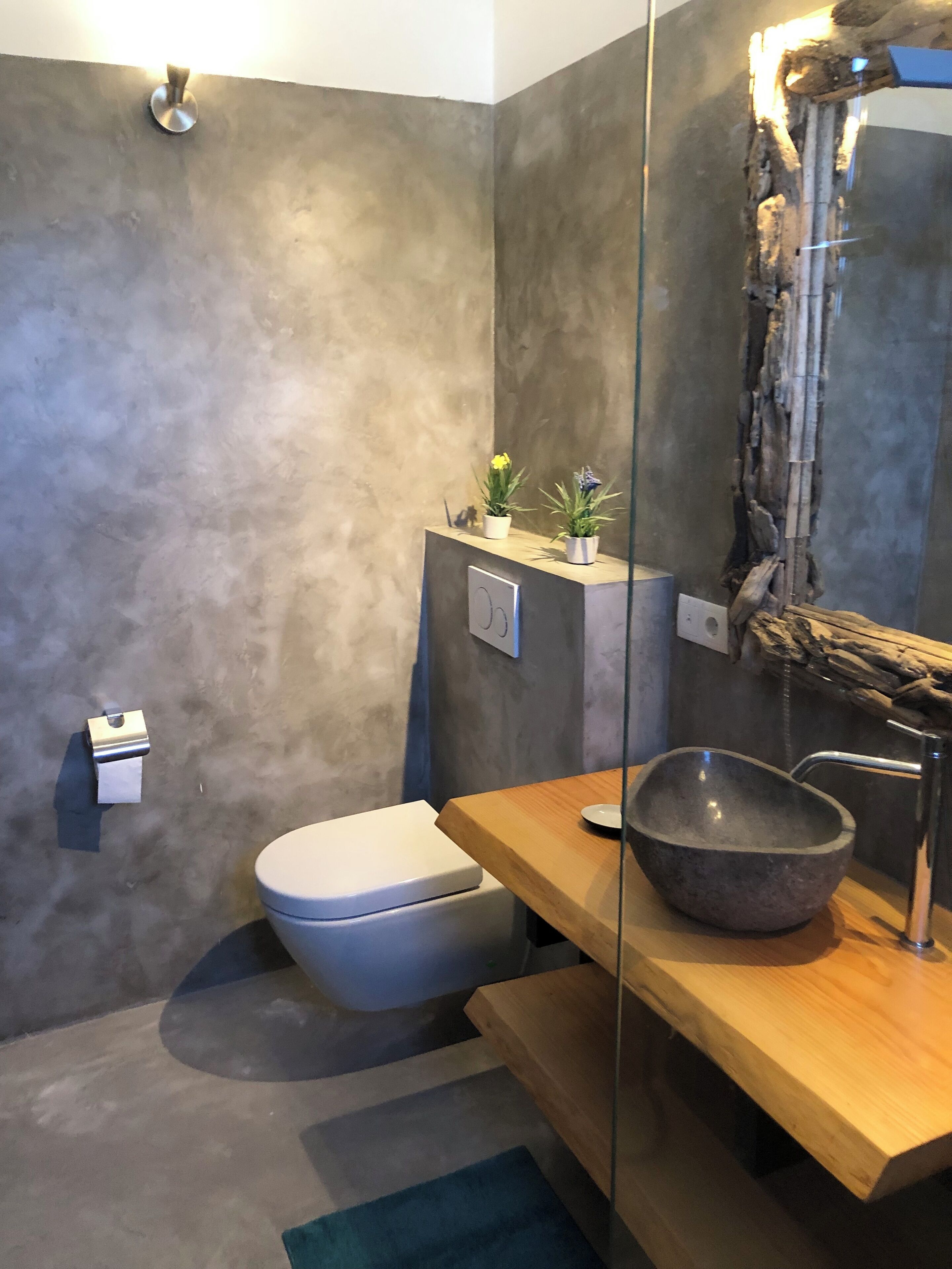 Een moderne badkamer met een betonnen muur, een wit toilet, een houten wastafelmeubel met een groot wastafelbakje, en een kleine potplant op het meubel.