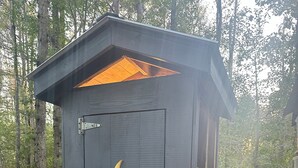 A small, dark-colored shed with a crescent moon cutout on the door, a slanted roof, and a metal latch.