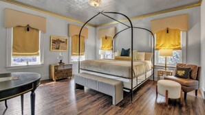 Un lit à baldaquin avec un banc, une table ronde et une chaise, dans une pièce au plancher en bois et dotée de grandes fenêtres ornées de rideaux jaunes.