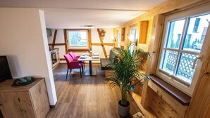 Et hyggeligt rum med trævægge, et spiseområde med bord og stole, en potteplante og et vindue med udsigt til et hegn og træer.