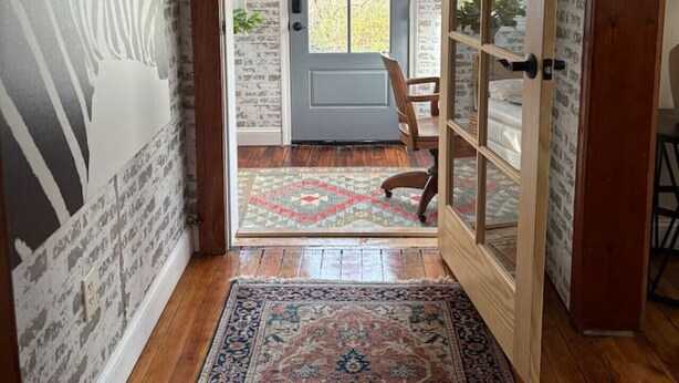 A hallway with a patterned wall, a wooden floor, a rug, a door with a wreath, and a bed visible through a glass door. - The Sun Filled Cozy Creamery (Plainville)