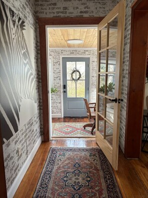 Un couloir avec un mur orné de motifs, un plancher en bois, un tapis, une porte décorée d’une guirlande, et un lit visible à travers une porte en verre.