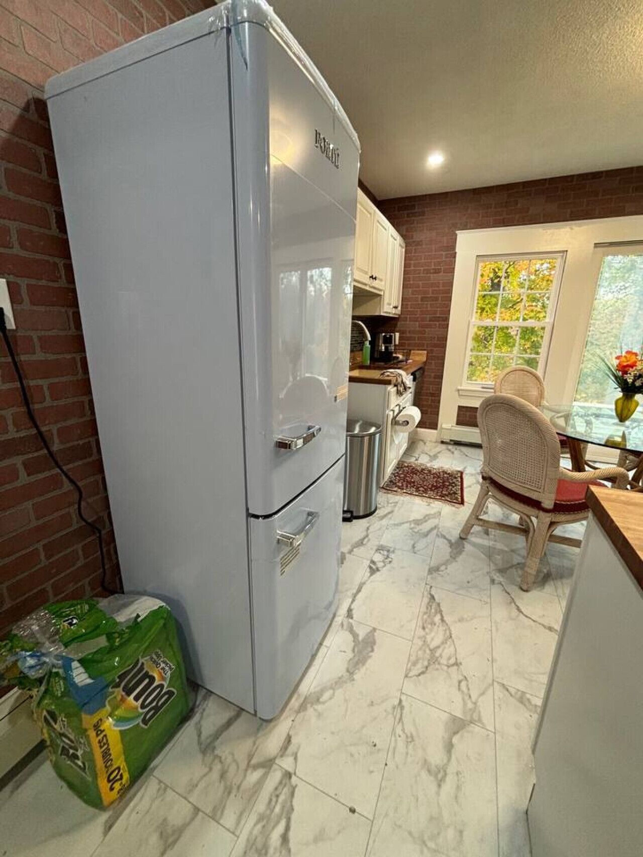 A compact kitchen with a white refrigerator, a dining area with a wicker chair, and a window with a view of trees.