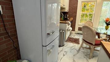 A compact kitchen with a white refrigerator, a dining area with a wicker chair, and a window with a view of trees.
