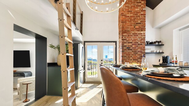 Een moderne keuken met een accentmuur van baksteen, een eethoek met een lange tafel en stoelen, en een uitzicht door de glazen deur.