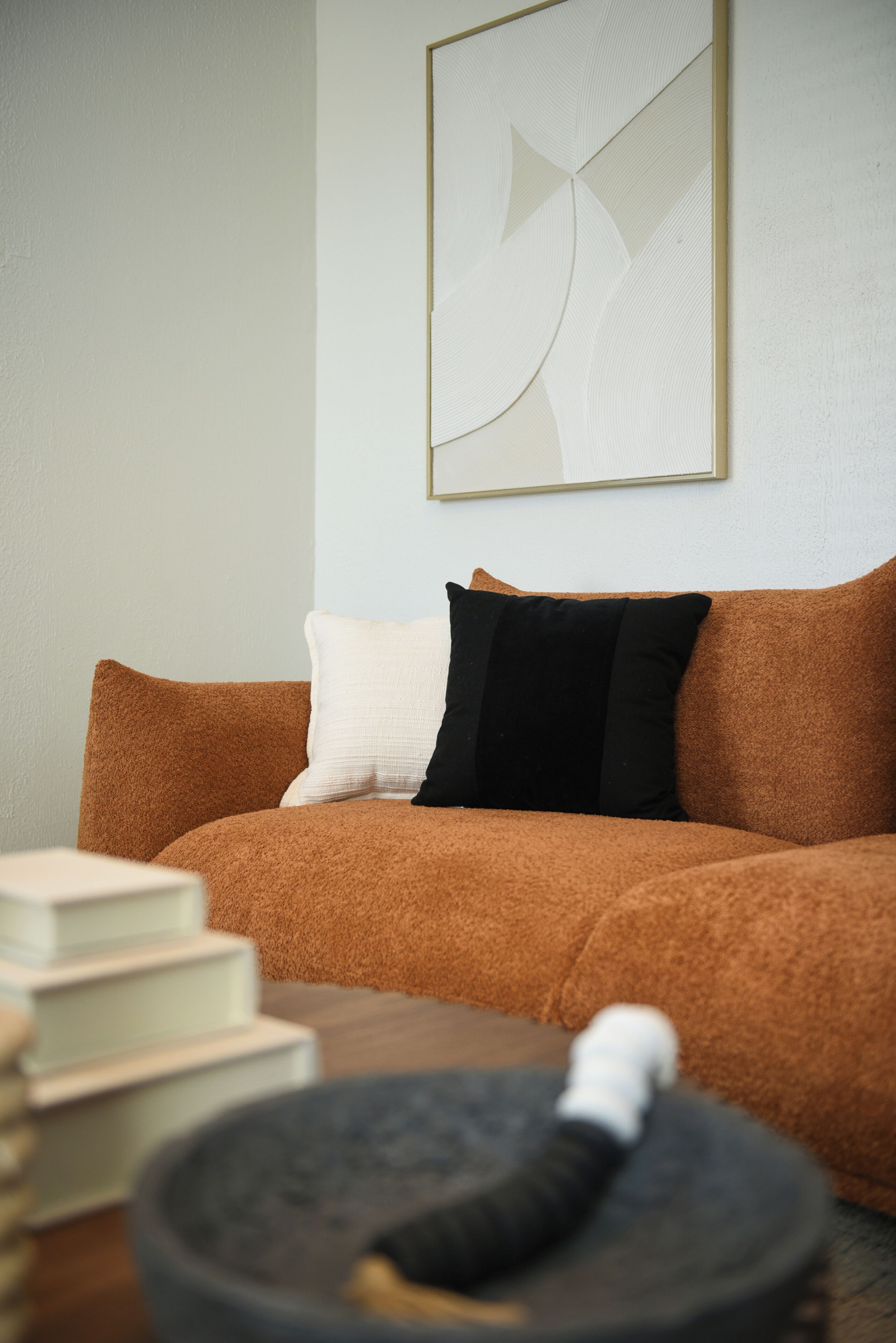 A living room with a brown sofa, a white and black pillow, a framed abstract artwork on the wall, and a round table with a black object on it.