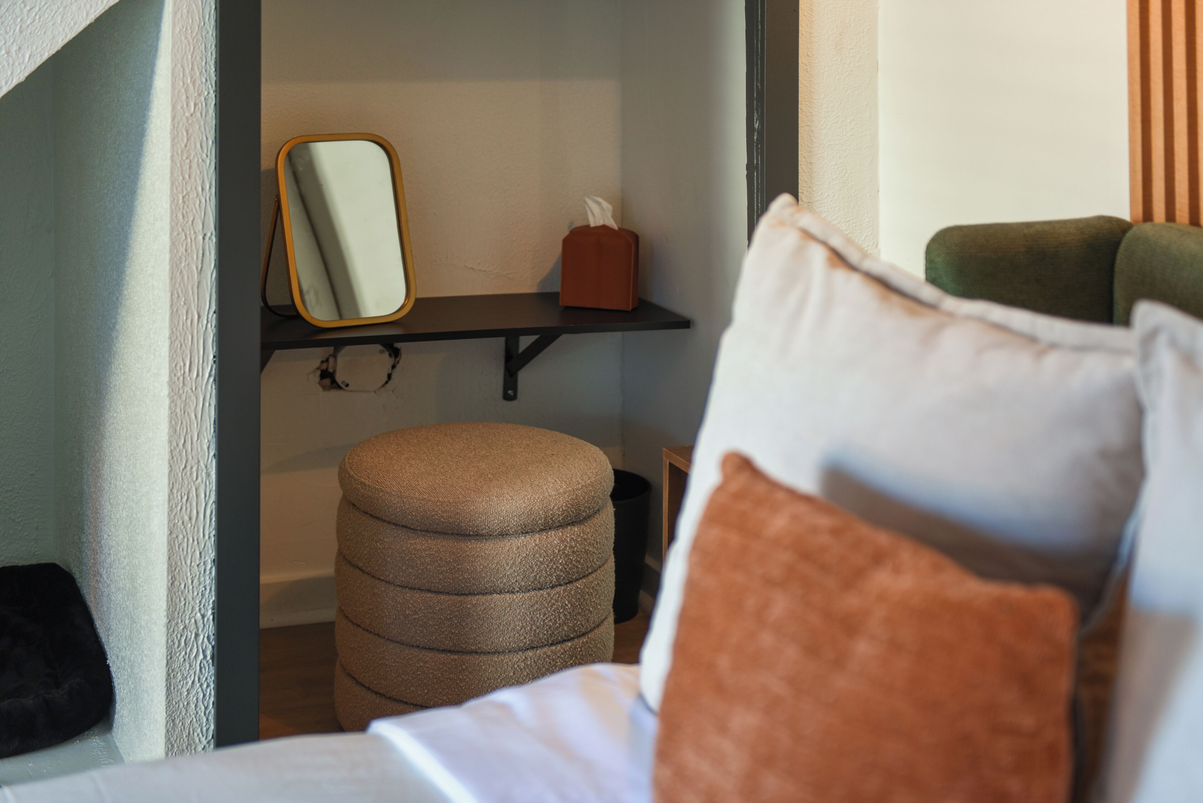Een slaapkamer met een bed, nachtkastje, spiegel, klein krukje en plank met zakdoosjes.