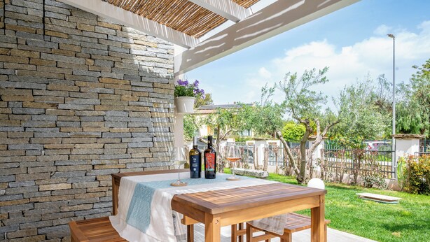 Eine Terrasse im Freien mit einer Steinmauer, einem Holztisch und -bänken, und Blick auf einen Garten sowie ein Haus im Hintergrund.