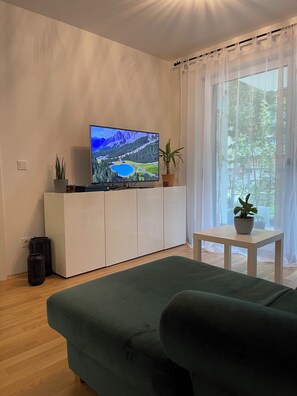 Un salon avec un téléviseur à écran plat posé sur un meuble blanc, un canapé vert, une petite table avec une plante et une fenêtre ornée de rideaux légers.