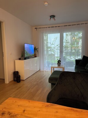 A modern living room with a flat-screen TV on a white cabinet, a green sofa, a wooden coffee table, and a large window with sheer curtains. - Luxury City Apartment in Munich (München)