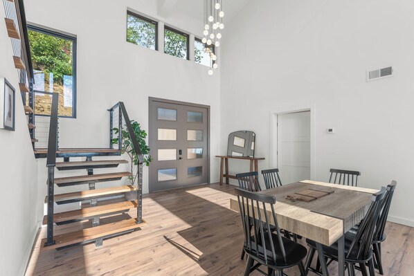 Ein großzügiger Essbereich mit Holztisch und -stühlen, einer Treppe mit Metallgeländern und großen Fenstern, die viel Tageslicht hereinlassen.