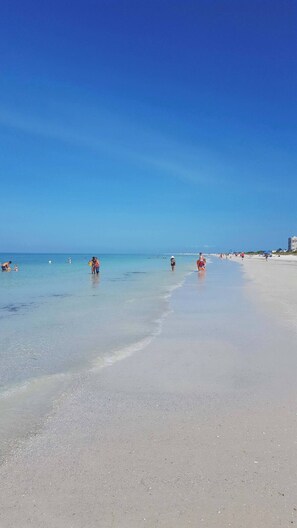 Una amplia playa arenosa con aguas azules y cristalinas, y algunas personas a lo lejos.