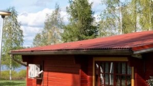 Un bâtiment en bois rouge, doté d’une fenêtre et d’une porte, entouré d’herbe et d’arbres.