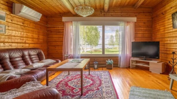 L’intérieur chaleureux d’un chalet en bois comprend un canapé en cuir, un téléviseur et une grande fenêtre donnant sur des arbres et un lac.