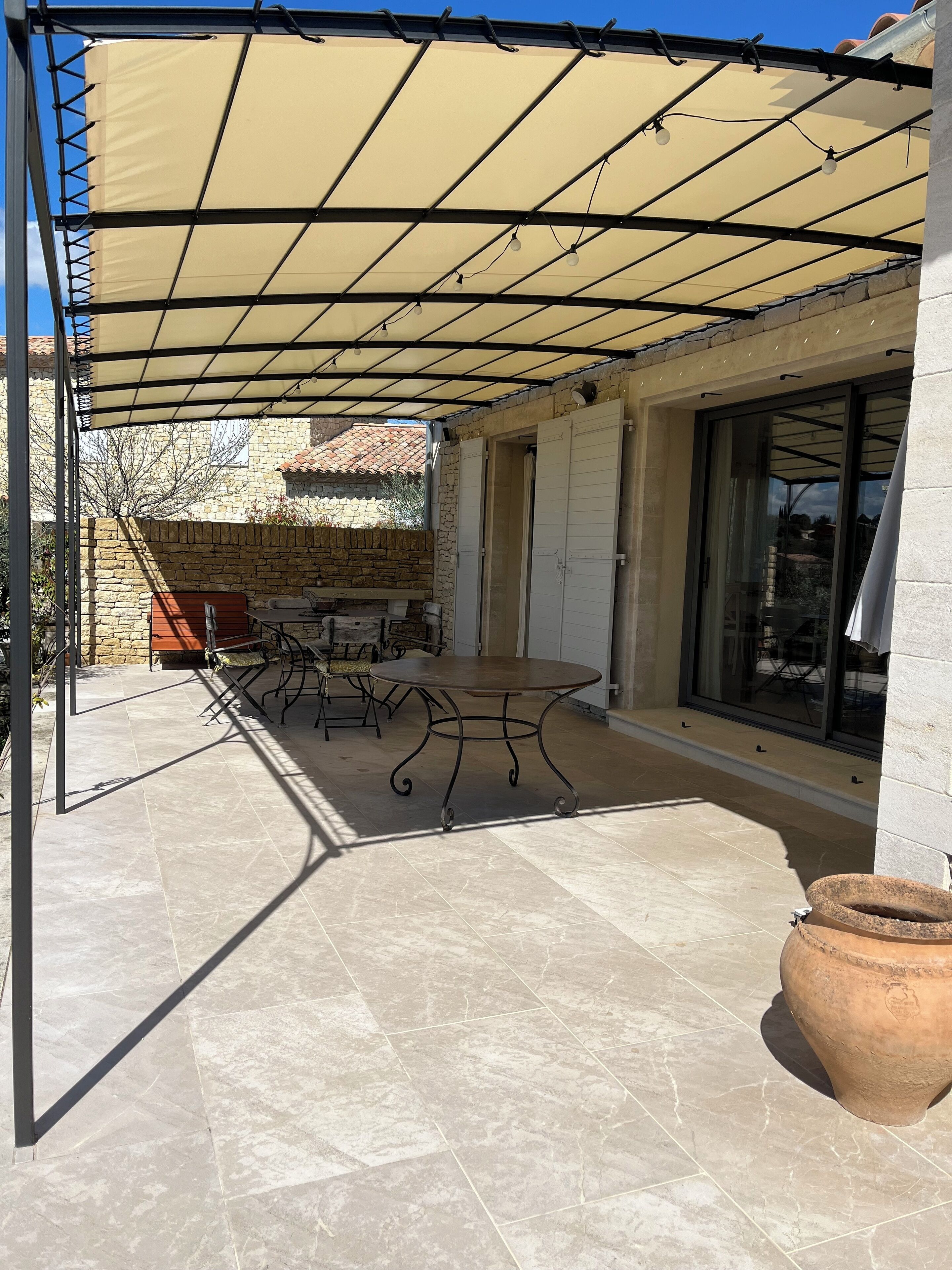 Un patio cubierto con una mesa y sillas, una planta grande en maceta y un muro de piedra al fondo.