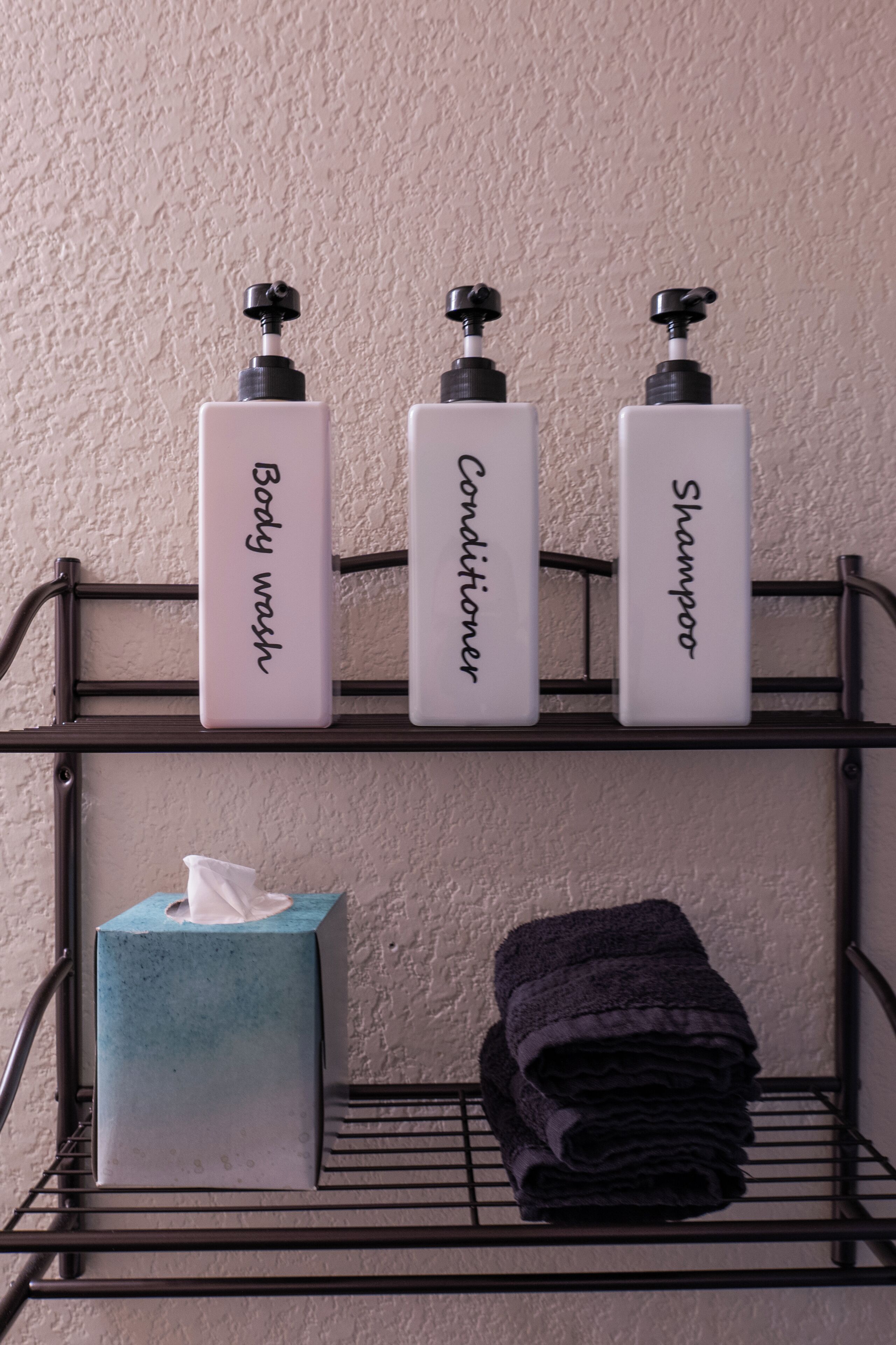 A bathroom shelf with three bottles labeled "Body wash," "Conditioner," and "Shampoo." Below, there is a tissue box and a stack of towels.