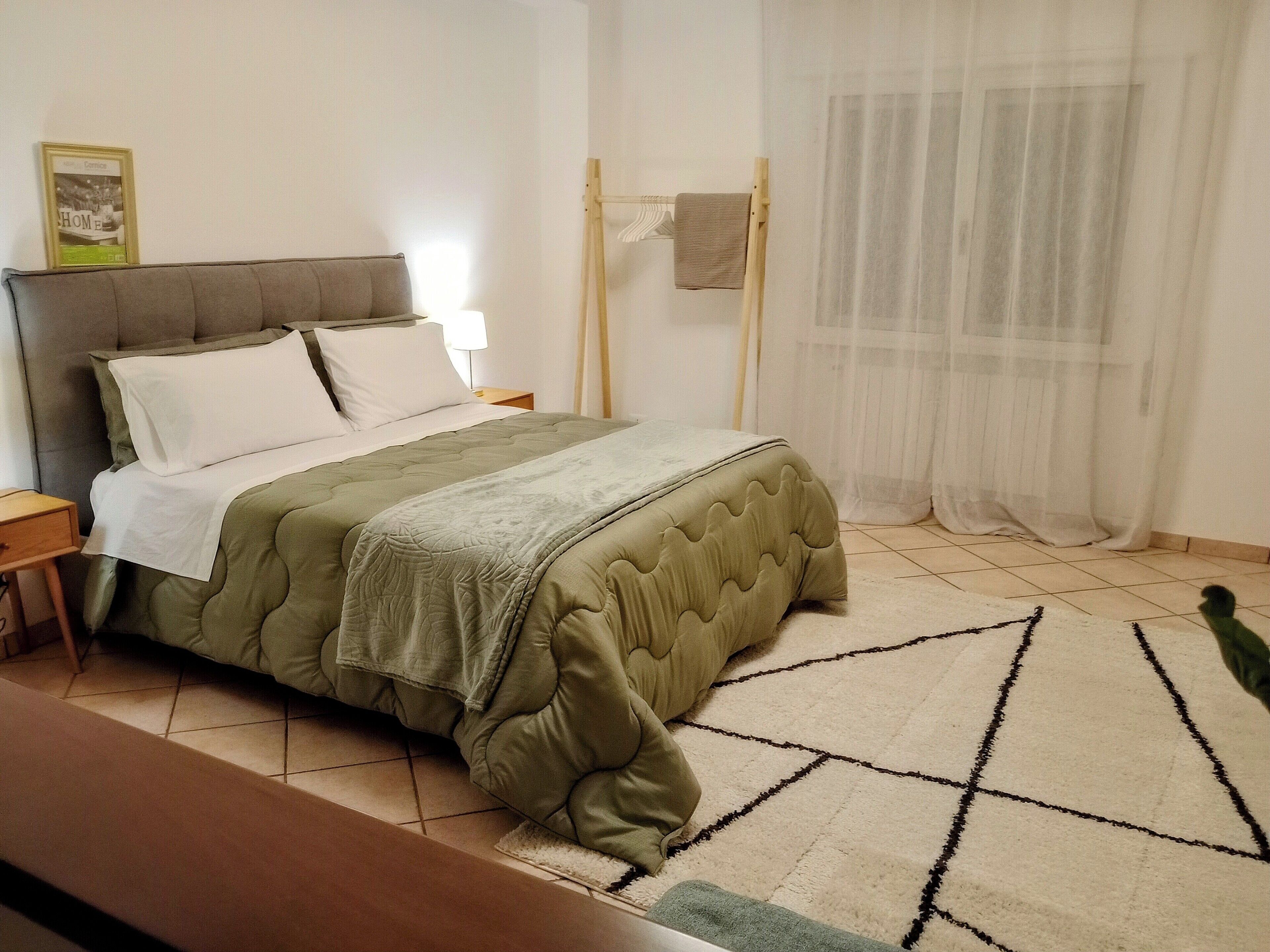 Ein ordentlich bezogenes Bett mit einer grünen Tagesdecke und weißen Kissen, ein Nachttisch aus Holz mit einer Lampe, ein gerahmtes Bild an der Wand und ein durchsichtiger weißer Vorhang.