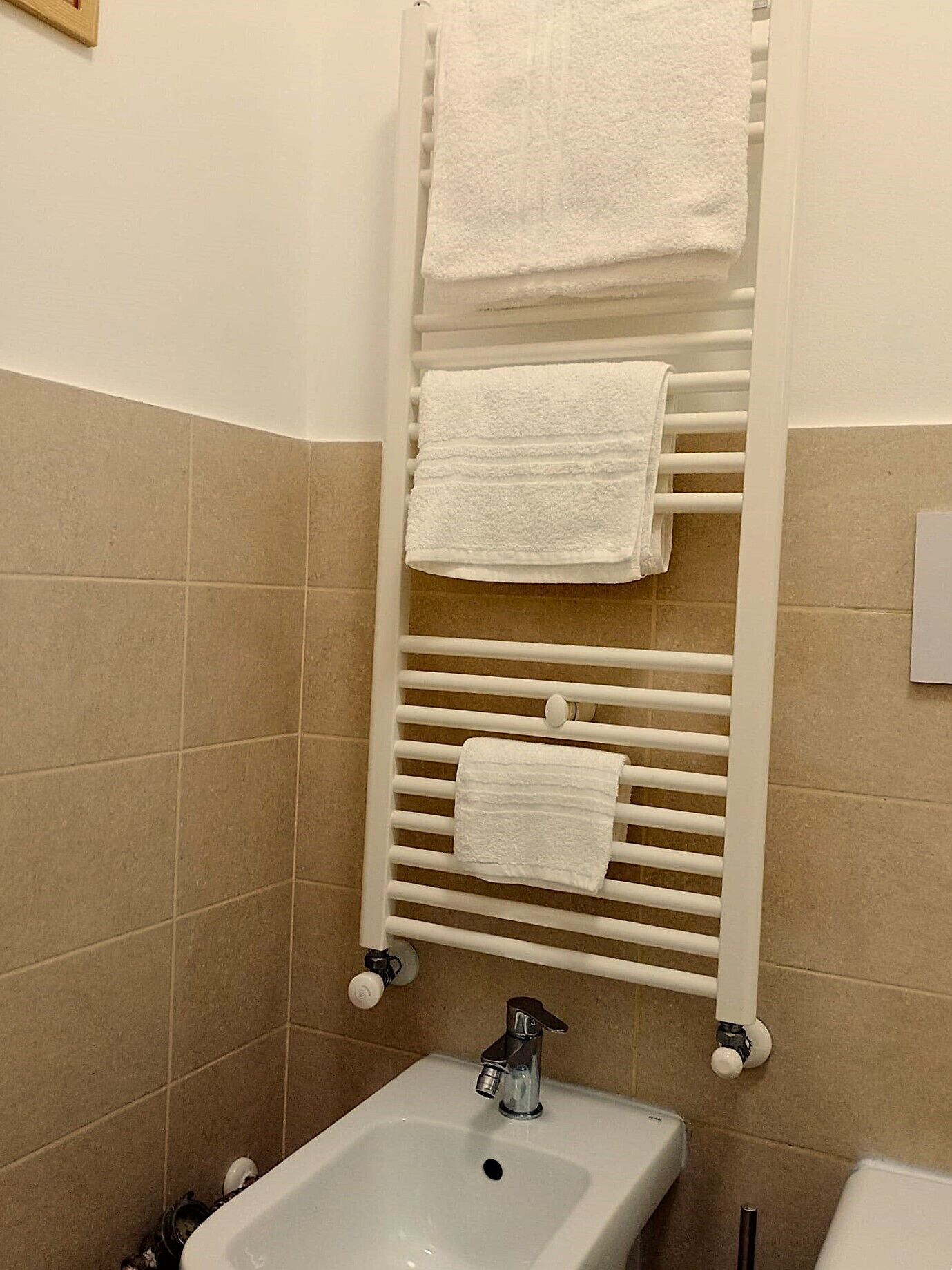 Un baño con un lavamanos blanco, un perchero con tres toallas blancas y paredes de cerámica en tonos beige.