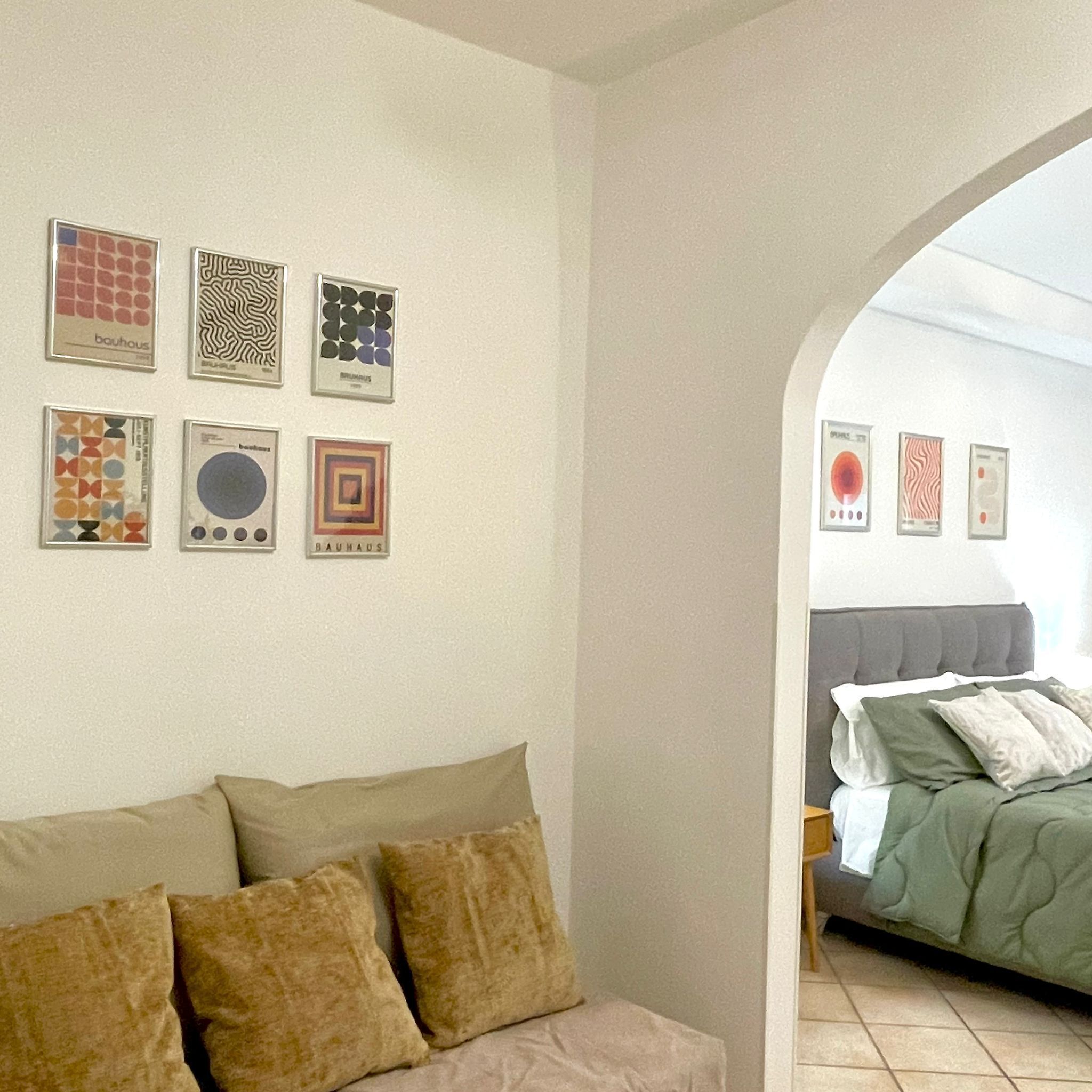 A room with a sofa and a bed, both with beige bedding. There are six framed artworks on the wall, each with different patterns and colors.
