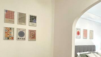 A room with a sofa and a bed, both with beige bedding. There are six framed artworks on the wall, each with different patterns and colors.