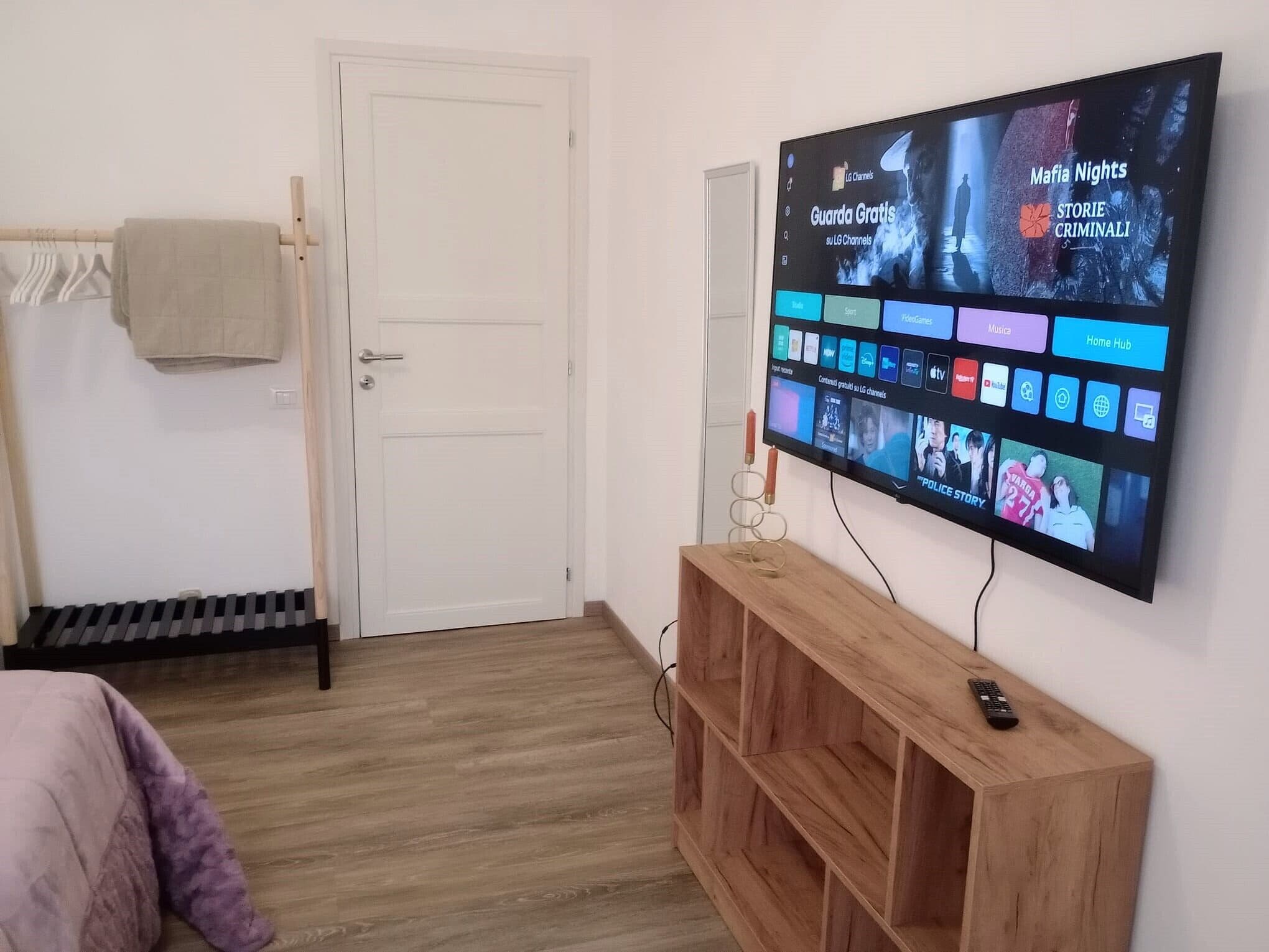 Una habitación con una televisión instalada en la pared mostrando varios canales, una mesita de madera con el control remoto, una puerta y un radiador.