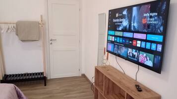 Una habitación con una televisión instalada en la pared mostrando varios canales, una mesita de madera con el control remoto, una puerta y un radiador.