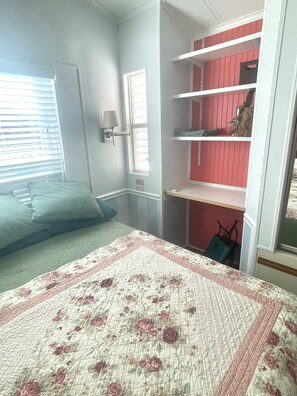 A bedroom with a bed, a window with blinds, a wall-mounted lamp, and a red shelving unit with various items.