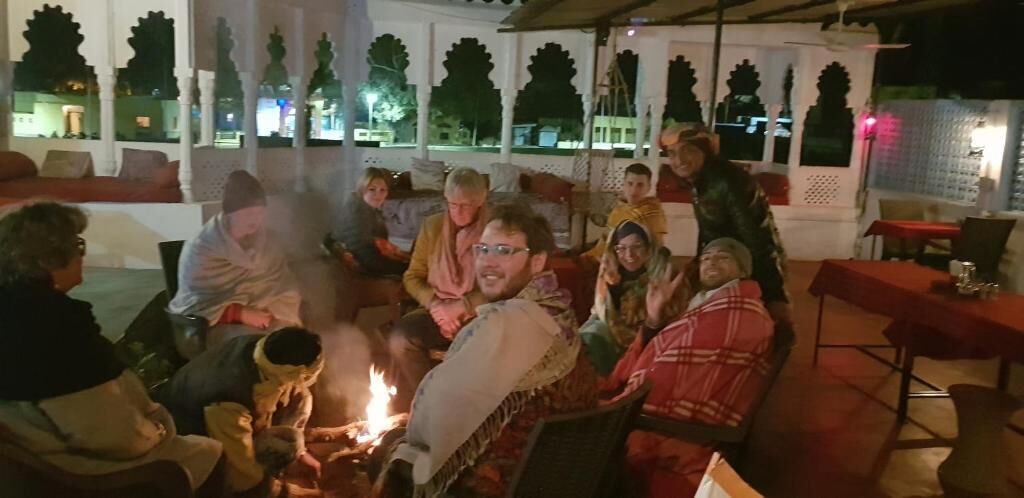 Eine Gruppe Menschen versammelt sich um ein Lagerfeuer in einer Freiluftumgebung, mit Blick auf Bäume und ein Gebäude im Hintergrund.