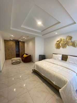A modern bedroom with a large bed, a wooden wardrobe, a seating area, and a wall-mounted decorative piece. - Green Court Apartments and Suites Lekki (Lekki)