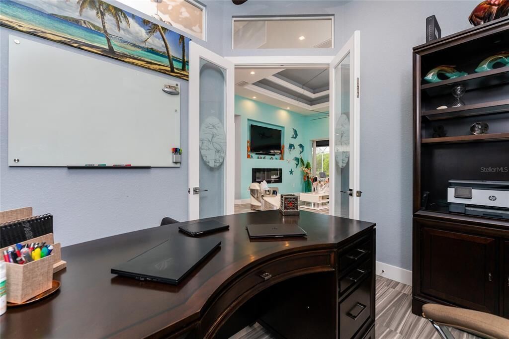 A room with a dark wooden desk, a whiteboard, and a bookshelf. There is a door leading to another room with a blue wall and a painting.