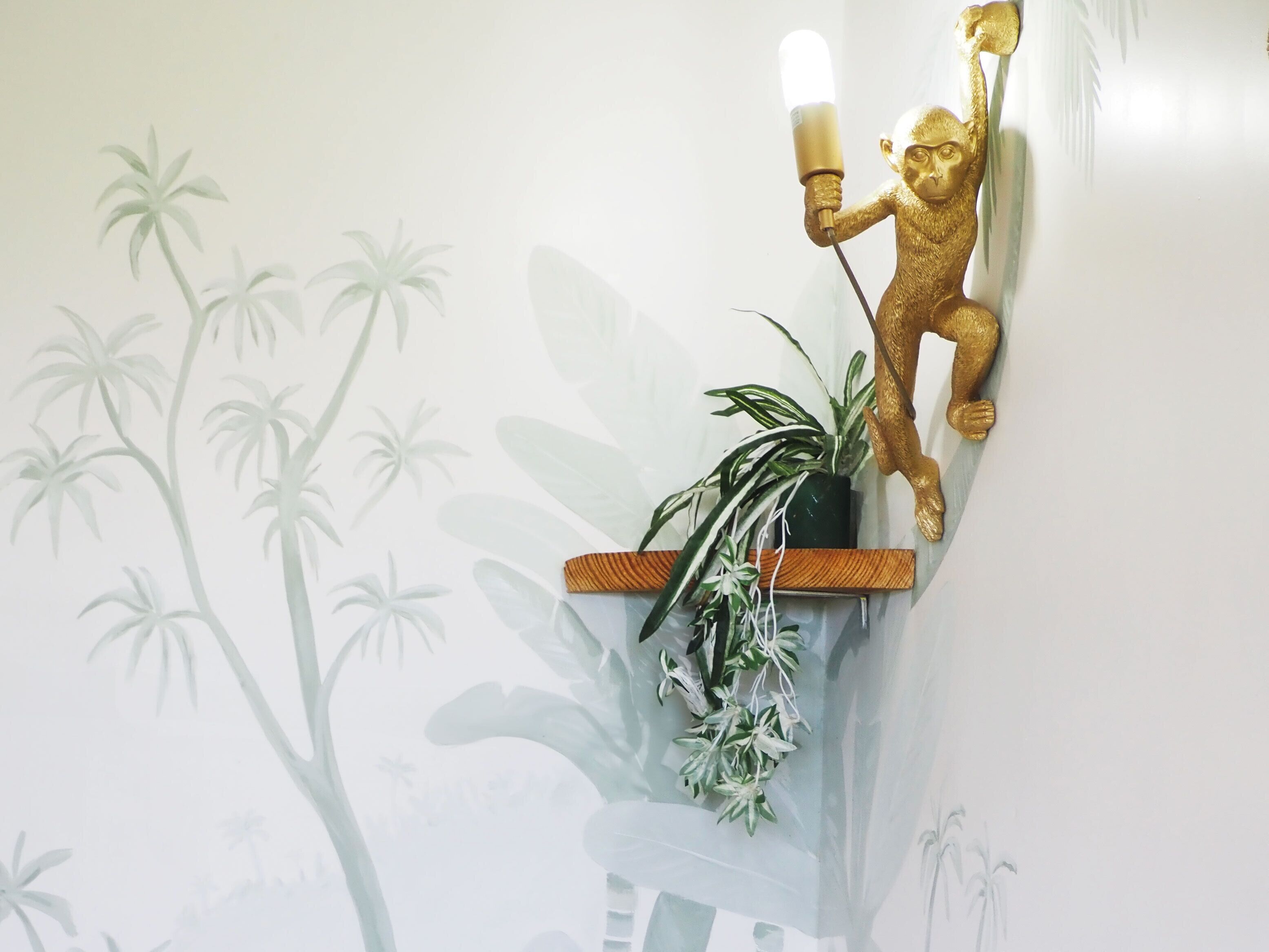A wall-mounted golden monkey sculpture holding a light fixture, a potted plant, and a wall with a tropical plant mural.