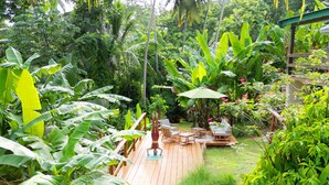 Una terraza de madera rodeada de abundante vegetación y un pequeño jardín con una mesa y sillas.