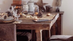 Uma mesa de madeira rústica posta com pratos, copos e velas, rodeada por cadeiras de couro marrom.