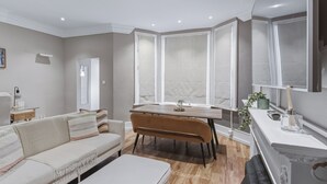 Een moderne woonkamer met een bank, een eettafel en een hocker. Er is een raam met zonwering en een deur die naar een andere kamer leidt.