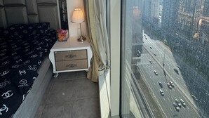 Habitación de hotel con cama, mesita de noche, lámpara y un ventanal con vistas a la ciudad.