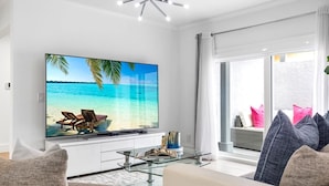 En moderne stue med et stort fladskærms-tv, der viser et strandbillede, et sofabord i glas og en sofa med puder.