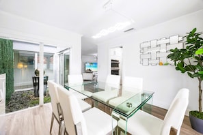 A modern dining area with a glass table and white chairs, a potted plant, and a wall-mounted decorative piece. - The Spectacular Miami Resort/5 Bedrooms/Htd Pool/MiniGolf/Playground/Baby Items (Miami)