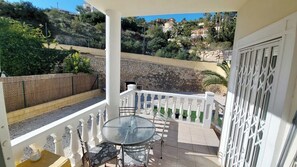 A balcony with a glass table and chairs, a white railing, and a view of a hillside with houses.