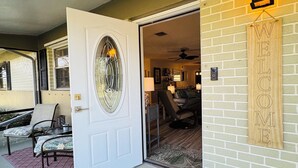 Une entrée d’habitation avec une porte blanche, une pancarte de bienvenue, un petit tapis orné d’un motif d’ancre, et un espace patio avec des chaises et une table.