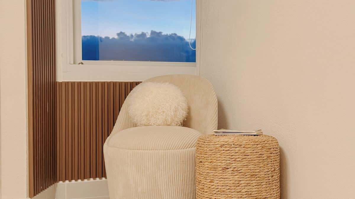 Una esquina de habitación con una ventana, una silla, un cojín y un taburete de mimbre.