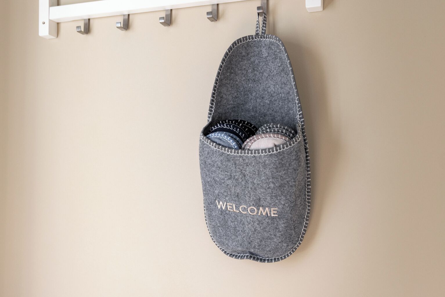 A grey felt slipper with 'WELCOME' embroidered on it, hanging on a white metal rack.