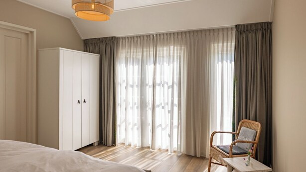 Un dormitorio con cama, armario, silla, mesa y un amplio ventanal con cortinas.
