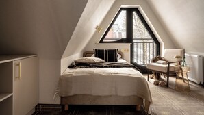 Une chambre avec un plafond en pente, un lit, une chaise, une table et une fenêtre donnant sur les bâtiments environnants.